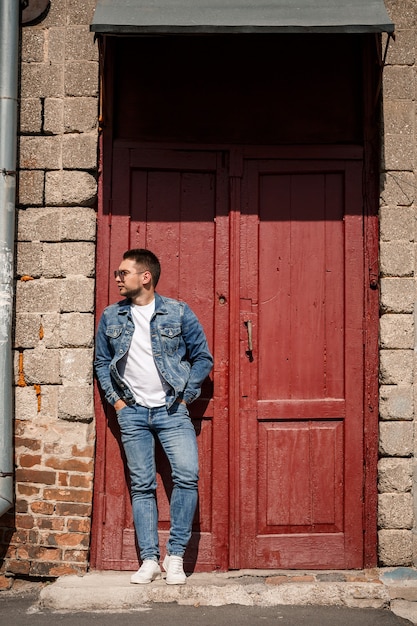 Un bell'uomo con la barba in giacca di jeans e pantaloni di jeans si trova vicino a porte vintage rosse. ragazzo cresciuto in stile denim