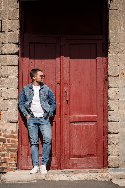 Handsome man with a beard in a denim jacket and denim pants stands near red vintage doors. Grown guy in denim style