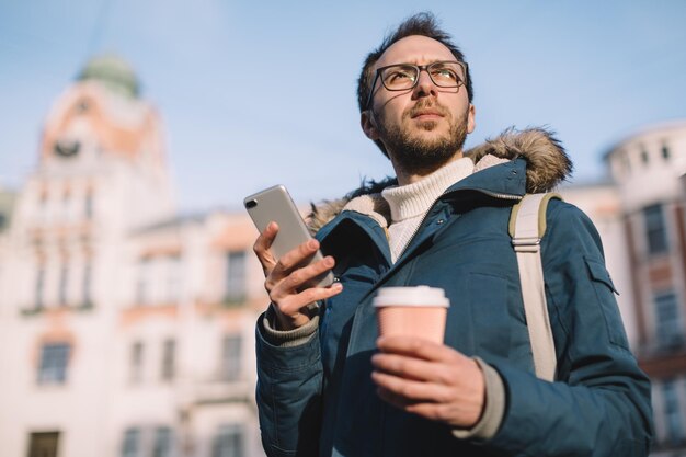 スマートフォンを持ち、コーヒーを飲む青いジャケットを着たひげと眼鏡をかけたハンサムな男性