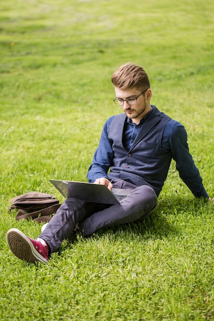 Handsome man with a beard is sitting on a lawn and working on a laptop