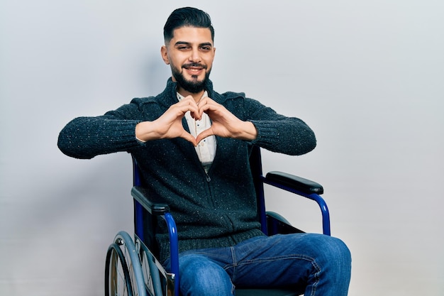 Handsome man with beard sitting on wheelchair smiling in love doing heart symbol shape with hands. romantic concept.