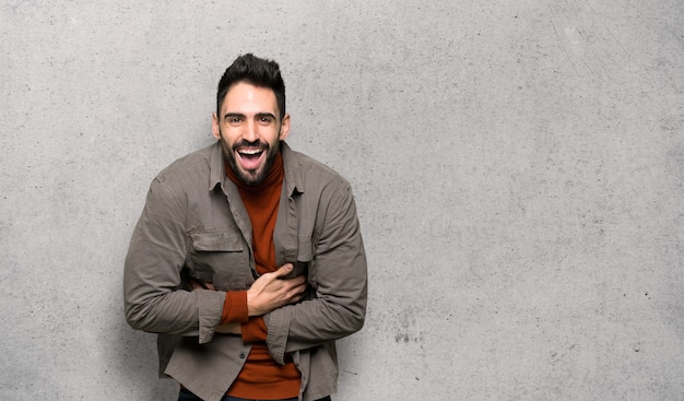Handsome man with beard smiling a lot while putting hands on chest over textured wall