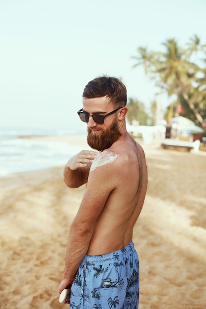 Uomo bello con la barba, in occhiali da sole, prendere il sole con crema solare corpo in estate.