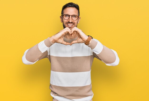 Handsome man with beard wearing casual clothes and glasses smiling in love doing heart symbol shape with hands. romantic concept.