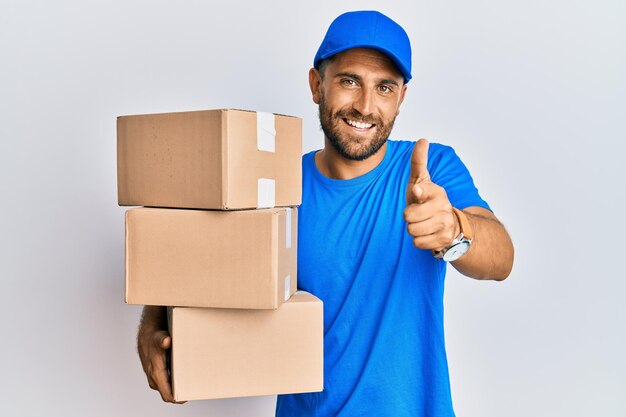 Handsome man with beard wearing courier uniform holding delivery packages pointing fingers to camera with happy and funny face. good energy and vibes.