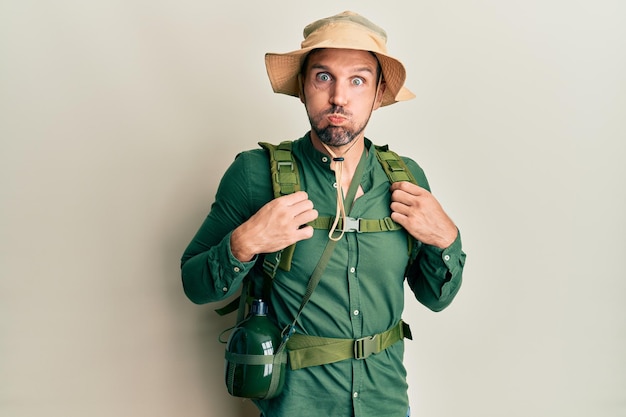 Handsome man with beard wearing explorer hat and backpack puffing cheeks with funny face. mouth inflated with air, crazy expression.
