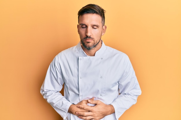 Handsome man with beard wearing professional cook uniform with hand on stomach because indigestion, painful illness feeling unwell. ache concept.