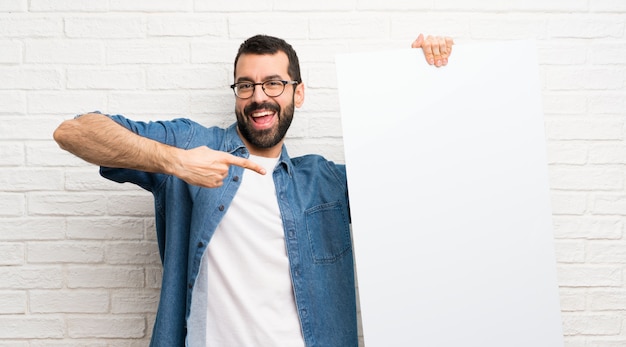 Handsome man with beard over white brick wall holding an empty placard