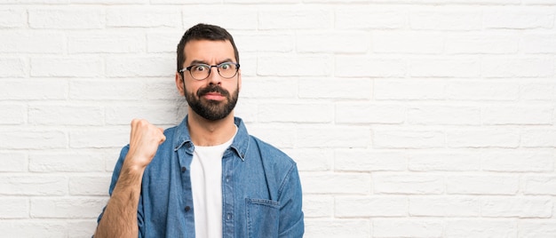 Handsome man with beard over white brick wall with angry gesture