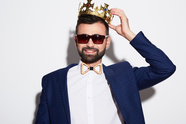 Photo handsome man with black hair and beard wearing white shirt, suit and sunglasses at white studio background, portrait, wearing crown.
