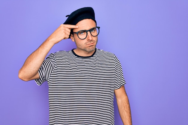 Handsome man with blue eyes wearing striped tshirt and french beret over purple background pointing unhappy to pimple on forehead ugly infection of blackhead Acne and skin problem