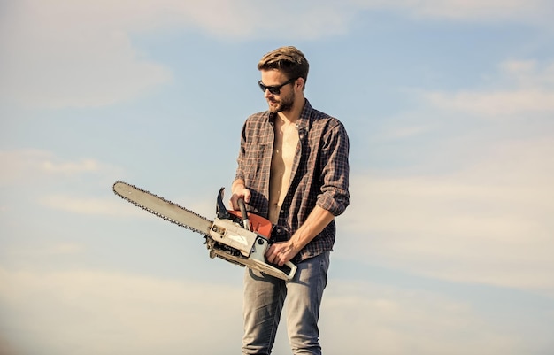 Photo handsome man with chainsaw blue sky background gardener lumberjack equipment lumberjack with chainsaw in his hands dangerous job feeling confident risky temper powerful chainsaw sharp blade