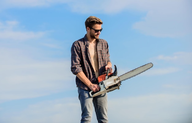 Handsome man with chainsaw blue sky background Gardener lumberjack equipment Lumberjack with chainsaw in his hands Dangerous job Feeling confident Risky temper Powerful chainsaw Sharp blade