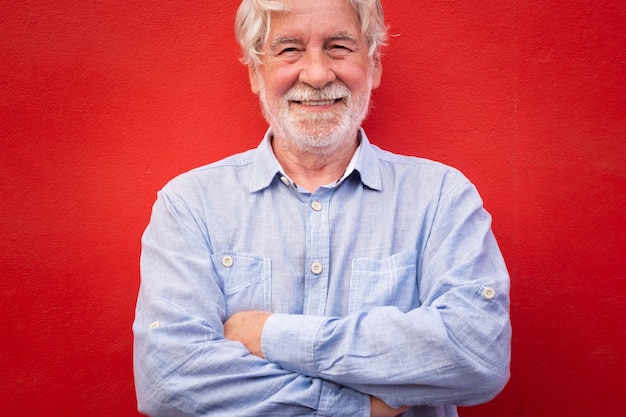 Handsome man with crossed arms standing on red background, smiling happily looking at camera, caucasian senior people with white beard and hair