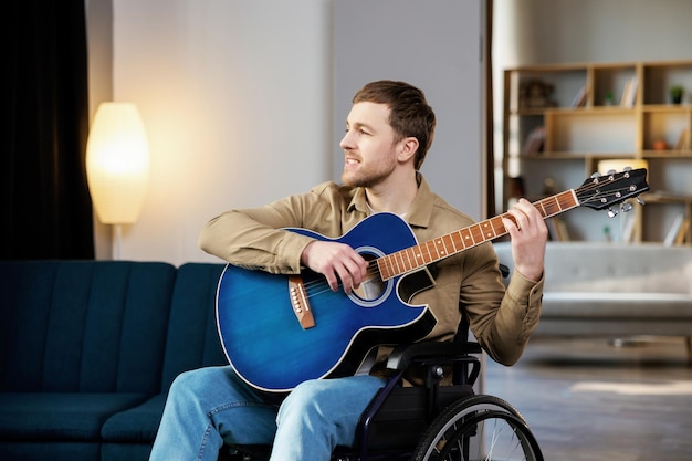 Handsome man with a disability sitting in wheelchair playing on guitar at home