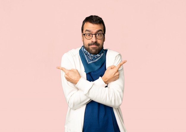 Handsome man with glasses pointing to the laterals having doubts on isolated pink background