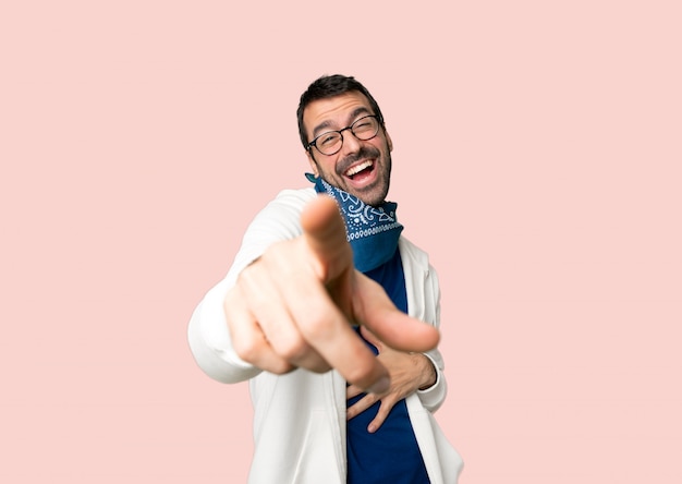 Handsome man with glasses pointing with finger at someone and laughing a lot on isolated pink background