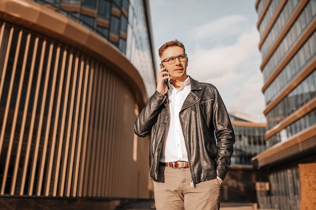 Bell'uomo con gli occhiali con uno smartphone sulla strada di una grande città. uomo d'affari che parla al telefono sullo sfondo urbano