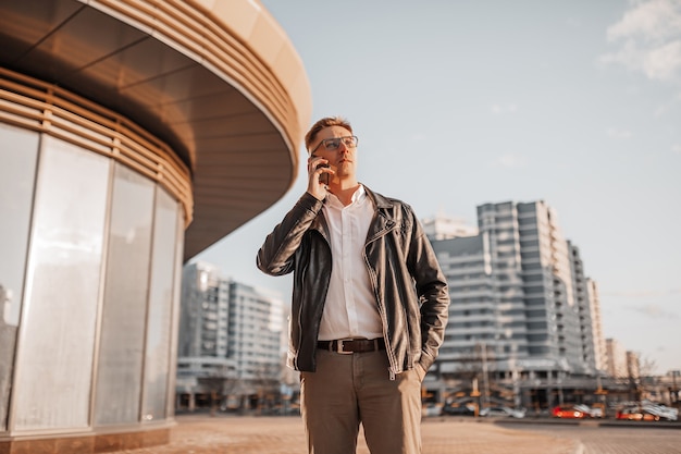 Handsome man with glasses with a smartphone on the street of a big city. Businessman talking on the phone on urban background