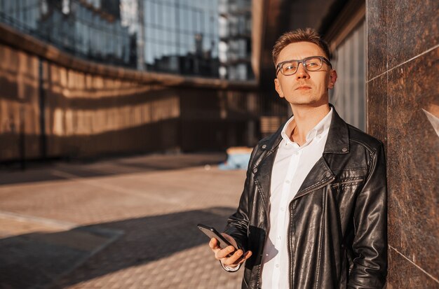 大都市の路上でスマートフォンを持った眼鏡をかけたハンサムな男。都市の背景に電話で話しているビジネスマン