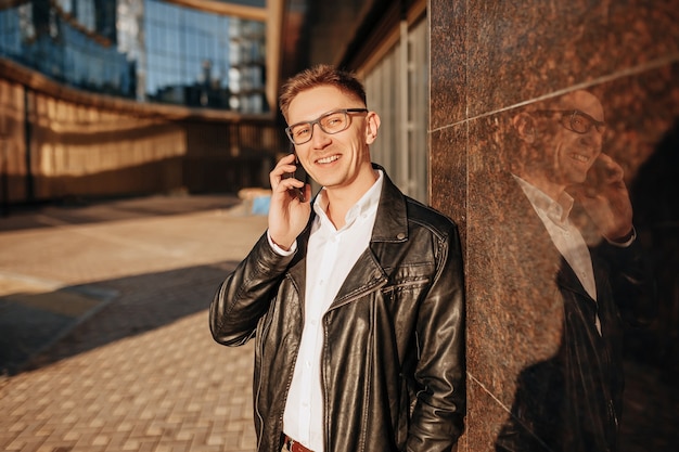 Handsome man with glasses with a smartphone on the street of a big city. Businessman talking on the phone on urban background