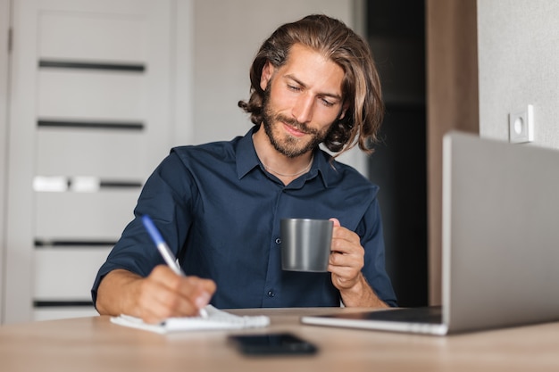 Handsome man with long hair writes in a notebook sitting at the table at home