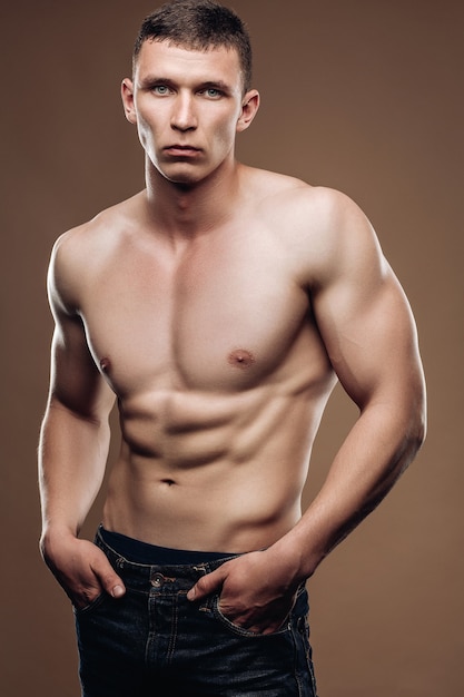 Handsome man with naked torso, muscular arms, press the cubes is in jeans, hands in pocket. Studio photos.