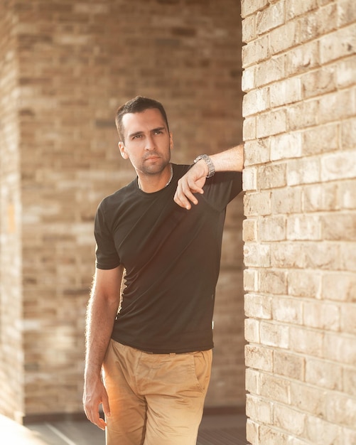 Handsome man with short bristles in a black Tshirt leans against a brick wall