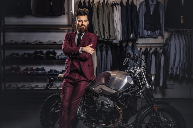 Handsome man with stylish beard and hair dressed in vintage red suit posing with crossed arms near retro sports motorbike at men's clothing store.