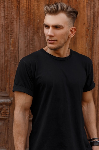Handsome man with stylish hairstyle in fashionable black mockup t-shirt stands near a wooden vintage door