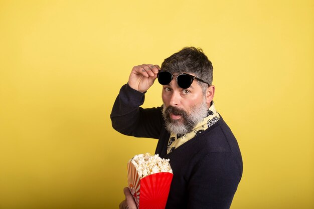 Handsome man with sunglasses in profile looking at camera holding a box full of popcorn on yellow background.