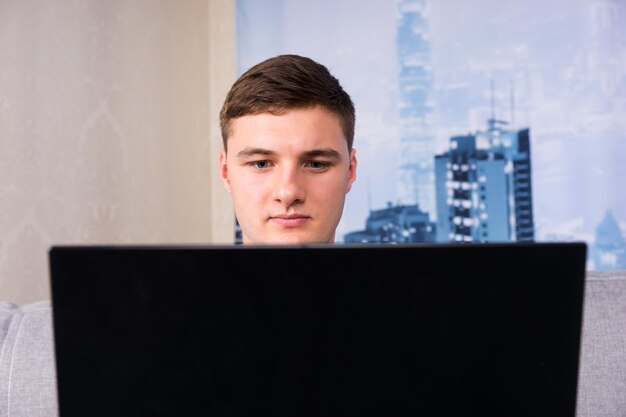 Handsome man working and sitting behind a laptop on a couch doing his business from home as he reads information on the screen