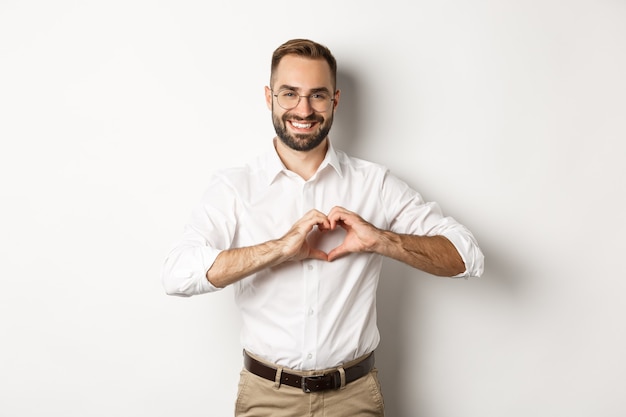 Handsome manage showing heart sign and smiling, I love you gesture, standing over white background.