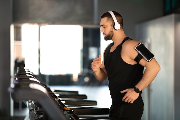 Photo handsome masculine man wearing wireless headphones running on treadmill at gym