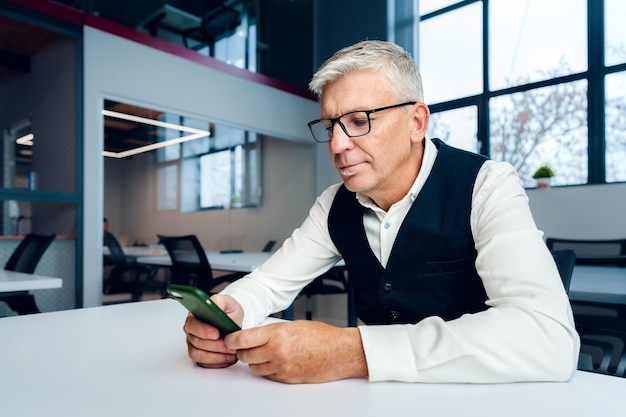 Uomo d'affari maturo bello usando il suo telefono cellulare al tavolo in ufficio