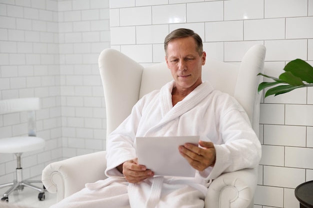 Handsome mature Caucasian man dressed in white bathrobe, with a blank paper sheet, sitting armchair in luxurious wellness spa centre