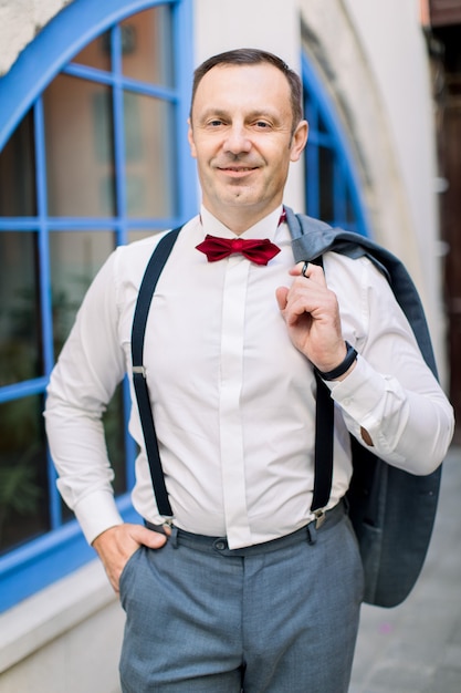 Handsome mature caucasian man groom in a white shirt with a red\
bow tie and in suspenders holds a jacket, posing in front of\
vintage building with big blue window outdoors in the city