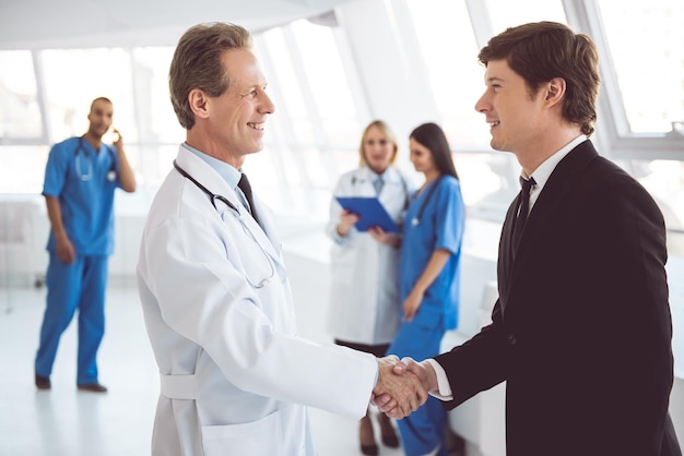 handsome mature doctor and young businessman are shaking hands and smiling while standing in the hospital hall