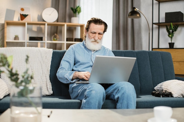 Handsome mature gentleman using laptop for video conversation sitting on sofa in living room at home