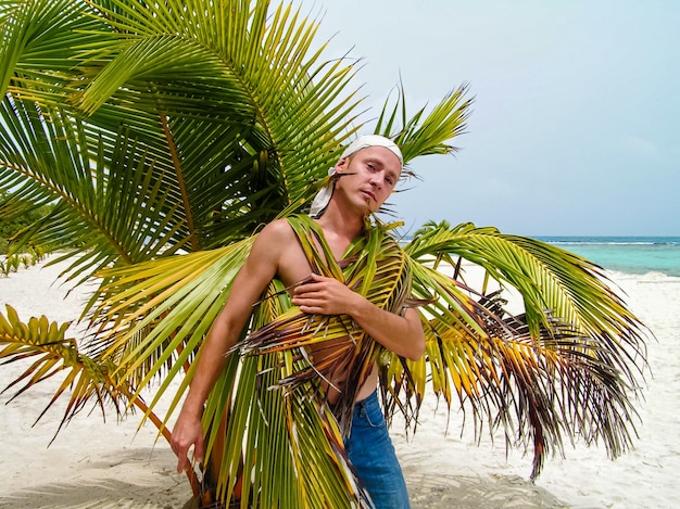 A handsome mature man stands topless under a palm tree on the sea beach, wrapped in leaves.