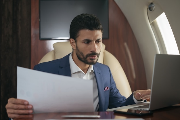 Photo handsome middle eastern businessman using laptop working with documents flying luxury jet plane