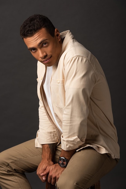 Photo handsome mixed race man in beige shirt sitting on chair on black background