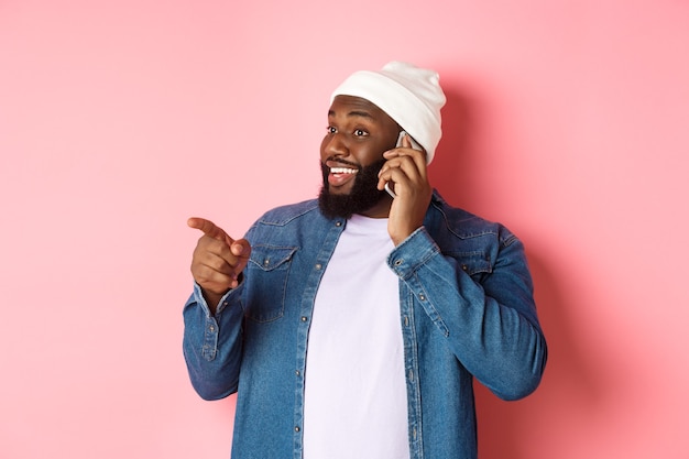 Handsome modern Black man talking on mobile phone, pointing left at person and smiling, standing over pink background