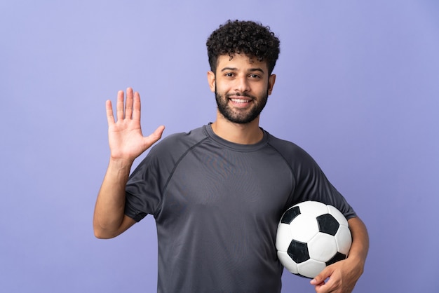 Handsome Moroccan young football player man over isolated on purple saluting with hand with happy expression