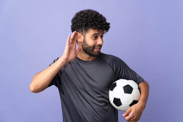 Handsome Moroccan young football player man over isolated on purple wall listening to something by putting hand on the ear