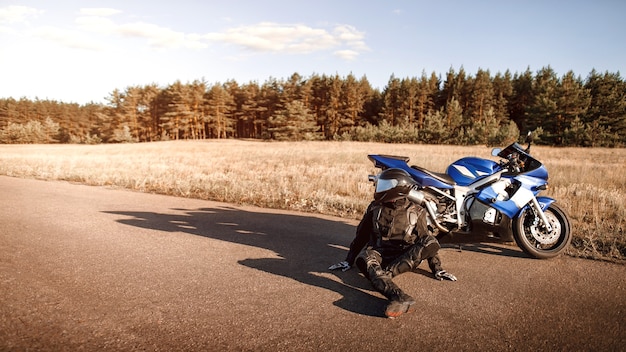 革のジャケットとヘルメットをかぶったハンサムなモーターサイクリストは、夕日の暖かい太陽光線の中で道路上のアスファルトのオートバイの隣に座っています