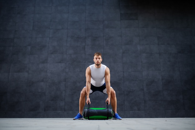 Handsome muscular bearded caucasian man in shorts and t-shirt lifting training bag while standing outdoor in front of gray wall.