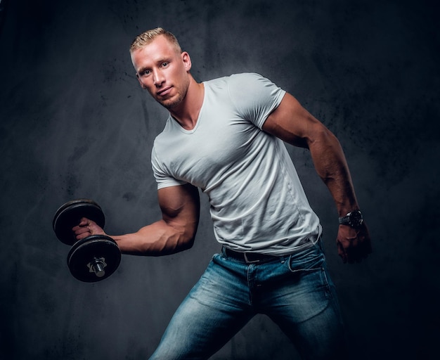 Handsome muscular blond male in a white t shirt holds dumbbell over grey artistic background.