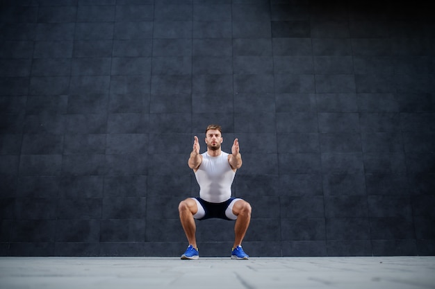 Handsome muscular caucasian man in shorts and t-shirt doing squatting exercise outdoors. In background is gray wall.