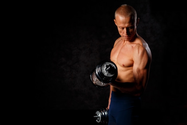 Handsome, muscular man with dumbbell over dark background.